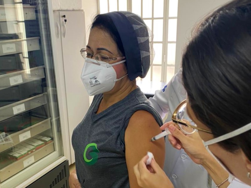 Foto de Rosália sendo vacinada. Ela está de toca e máscara de proteção e uma profissional da saúde aplica a vacina em seu braço
